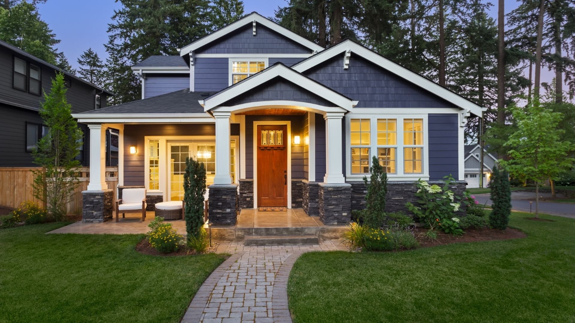 Modern blue and white house with lit porch, red door, manicured lawn and landscaping at dusk.