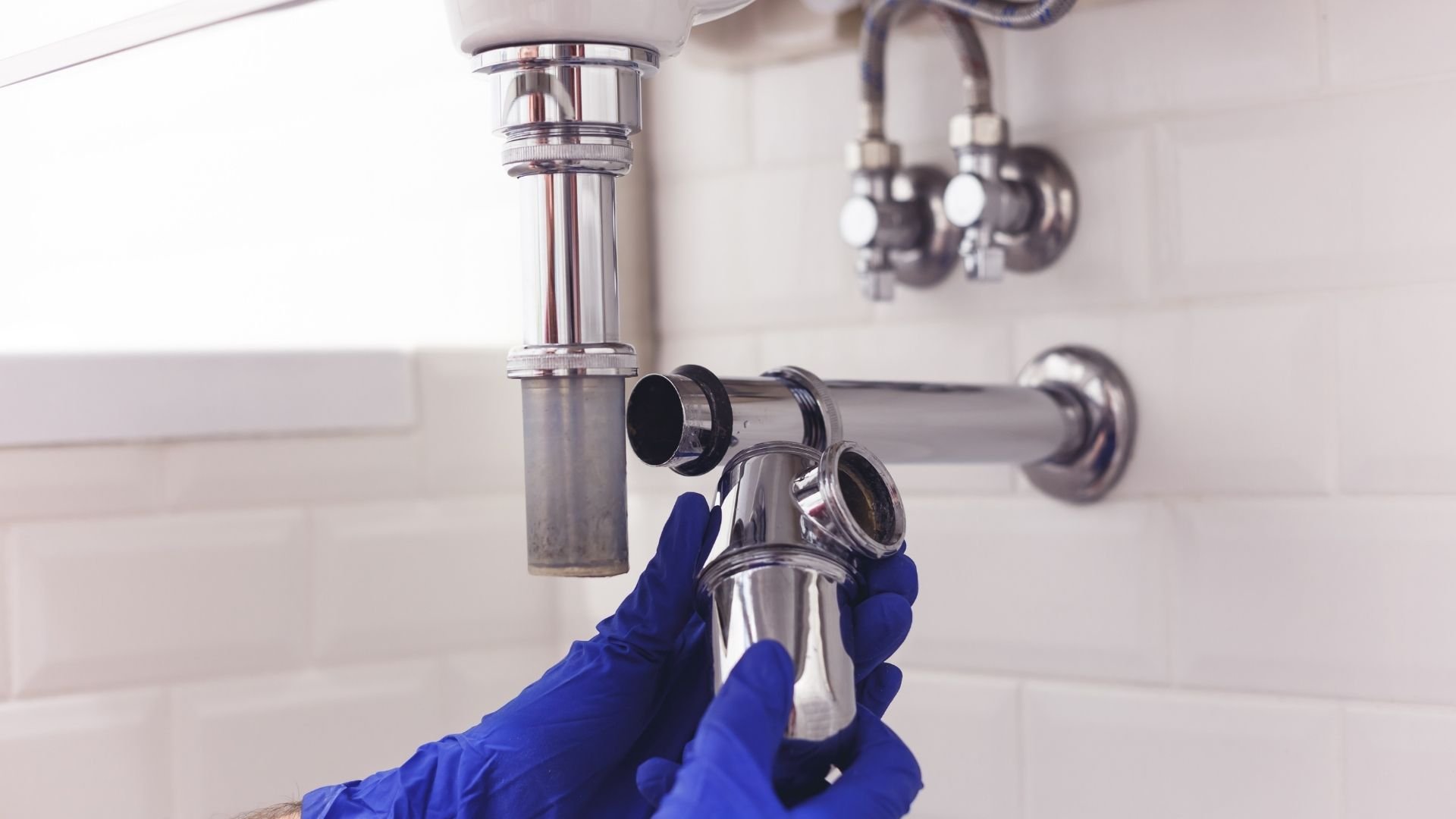 Plumber in blue gloves installing chrome sink pipe and P-trap under bathroom sink